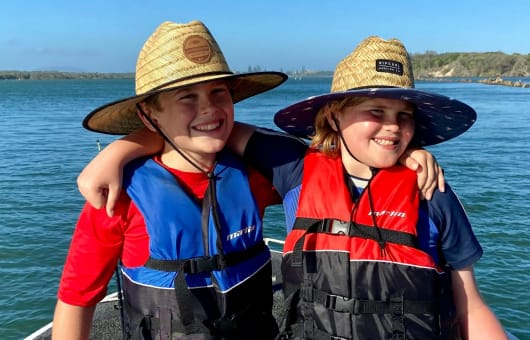 Two kids with arms around each other, wearing life jackets and straw hats.